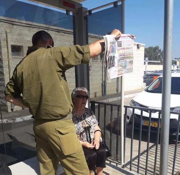 An IDF Soldier Provides An Elderly Woman At A Bus Stop With Shade